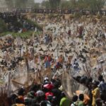 Dozens of Nigerian men in colorful shorts sprint and splash into the muddy Matawalle River at the 2026 Argungu Fishing Festival, holding large handmade nets, while spectators watch from the riverbank under a hazy sky.