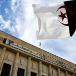 Contemporary glass skyscraper reflecting warm orange and pink sunset colors in its mirrored façade, set against a bustling city skyline