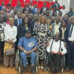 Young adult woman with long dark hair wearing a denim jacket, standing outdoors on a bright sunny day, smiling directly at the camera