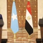 A vibrant orange and pink sunset reflected on a mirror-like lake, framed by dark mountain silhouettes and a small solitary boat in the distance
