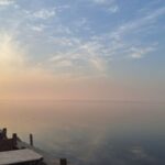 A rust-colored drainage pipe jutting from a dirt bank discharges dark wastewater directly into the calm blue-green surface of Lake Qarun near Shakshuk Village, Fayoum Governorate.
